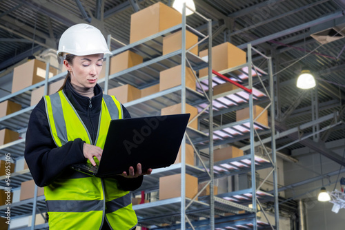 Storekeeper woman. Girl with laptop and shelving. Warehouse with boxes behind storekeeper. Storekeeper manages warehouse via laptop. Warehouse accounting software. Woman in front of blurred storage