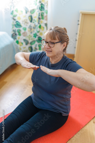 Active senior woman doing abs exercises on yoga mat at home and smiling at camera. Sporty mature lady working out in living room interior. Strength training concept