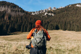 Back view of stylish woman wearing green backpack and red hat looking at mountain view while relaxing in nature. Travel and wanderlust concept. Amazing chill moment