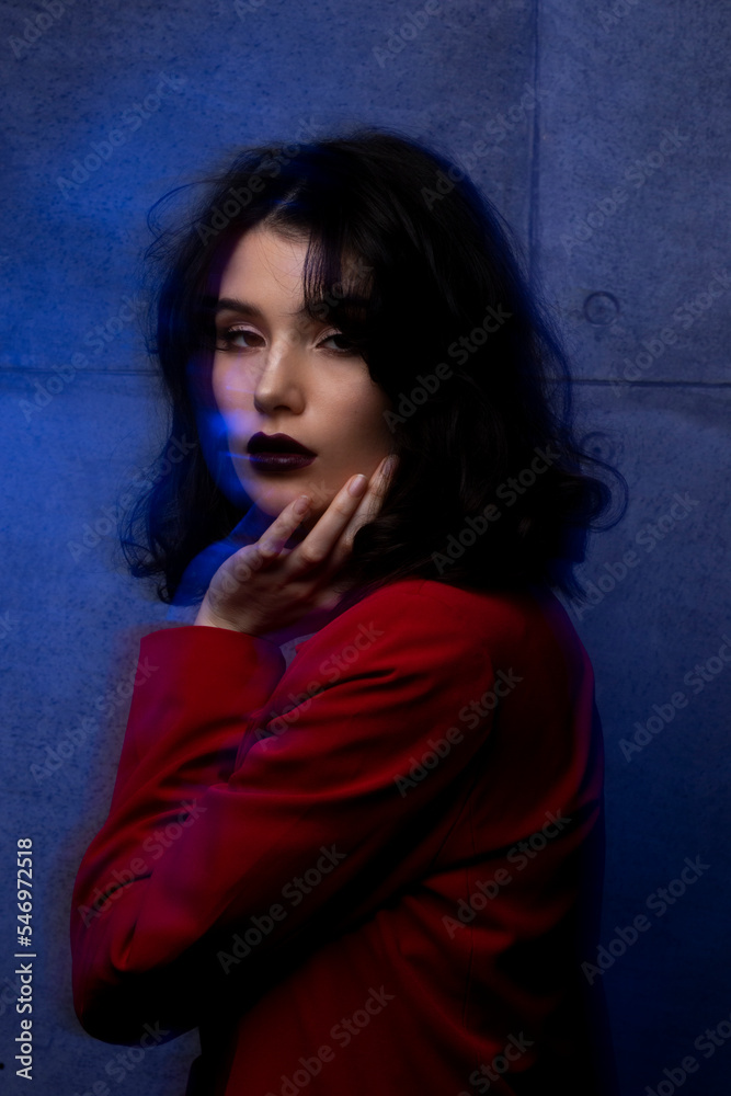 Portrait of young woman with makeup and hairstyle, wear red suit, looking at camera sensual, blue neon studio light.