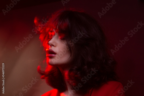 Closeup view of young woman with makeup and hairstyle, wear red suit, with closed eyes, red studio light and effects.
