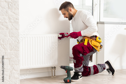 Man in work overalls using wrench while installing heating radiator in room. Young plumber installing heating system in apartment. Concept of radiator installation, plumbing works and home renovation.