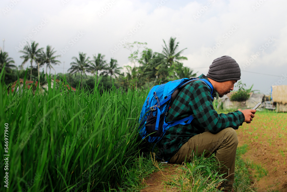 Young Asian man sitting next to a rice field in Indonesia while playing with his phone.