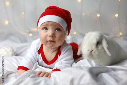 Wallpaper Mural Baby and white rabbit lie in bed. Happy litlle kid in red Christmas hat with rabbit. Child's Christmas gift in bed. Boy in a Santa hat with a rabbit. Funny pet. Christmas lights are on Torontodigital.ca