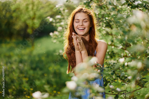 Woman with flying hair in spring stands in the orchard of a blooming apple tree and smiling with teeth stroking herself with her hands and embracing, in a blue dress, happiness sunset