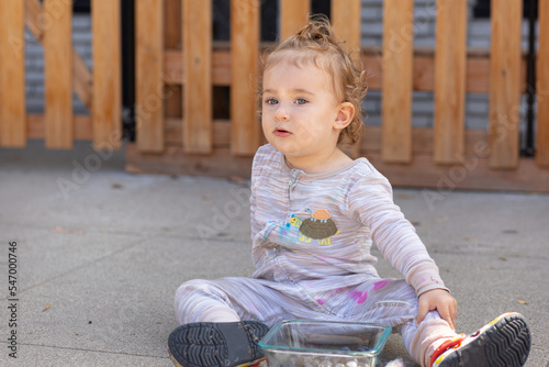 Portrait of a Toddler Girl. A baby is in front of a fence. Curly girl sits on the ground and she eats blueberry 