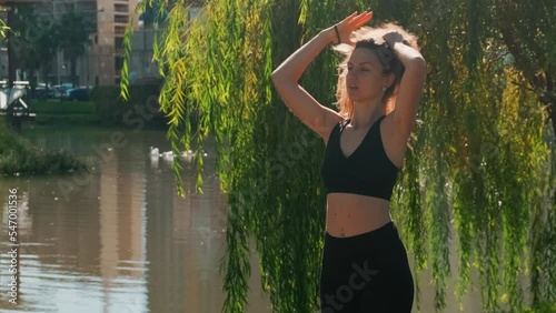 Woman straightens her long curly chic hair with her hands and collects it in ponytail. Sportswoman on the background of urban pond on which couple of swans swims. photo