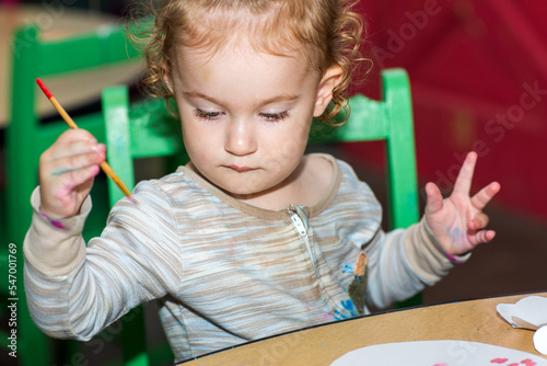 Toddler girl during art project. Kid holds paintbrush. Baby paints with brush on a paper. A girl paints a picture