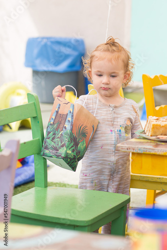 Portrait of a Toddler Girl. A baby holds a wooden spoon and a paper bag, and she eats a cake. Curly girl is next to a chair on Happy Birthday party 