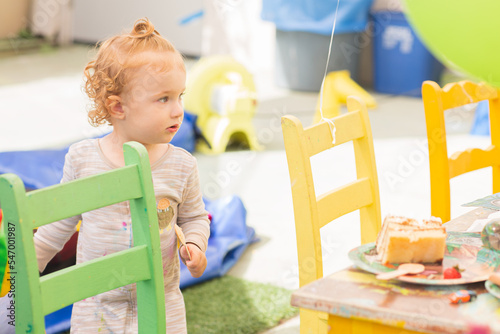 Portrait of a Toddler Girl. A baby holds a wooden spoon and a paper bag, and she eats a cake. Curly girl is next to a chair on Happy Birthday party 