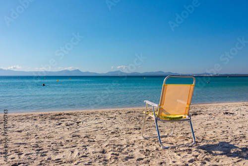 Fototapeta Naklejka Na Ścianę i Meble -  Playa de Alcudia (Mallorca). Silla de playa amarilla en la arena mientras alguien se baña en las aguas crsitalinas de la playa. Islas Baleares, España.