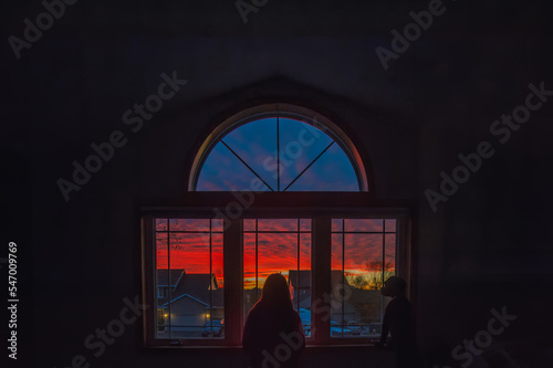 A girl and her dog look out a window at a colorful sunset over suburban houses