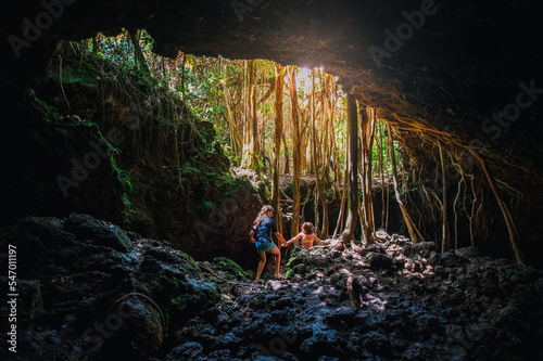 Two children make their way to the exit of a rocky cave with tree roots hanging down.