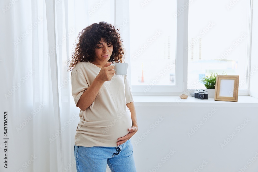 A pregnant woman stands at the window with a mug of warm water and tea and enjoys the view in a home t-shirt, the happiness of motherhood A pregnant woman stands at the window with a mug of warm water and tea and enjoys the view in a home t-shirt, the happiness of motherhood