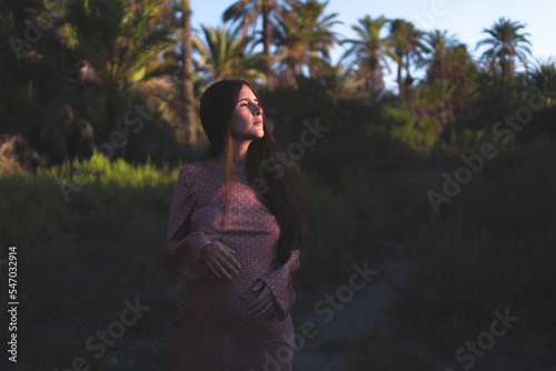 young pregnant woman relaxes in the golden light of sunset in a green natural place of plants and palm trees