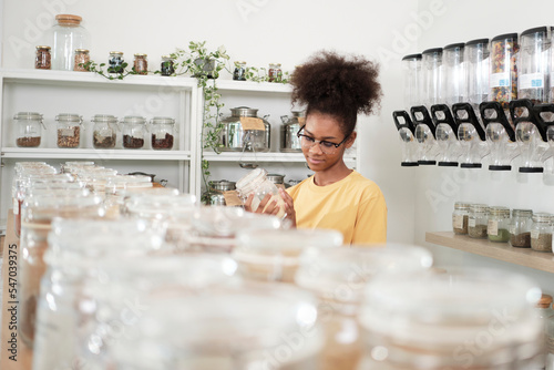 Young African American woman is choosing and shopping for organic products in refill store with reusable container, zero-waste grocery, and plastic-free, environment-friendly, sustainable lifestyles.