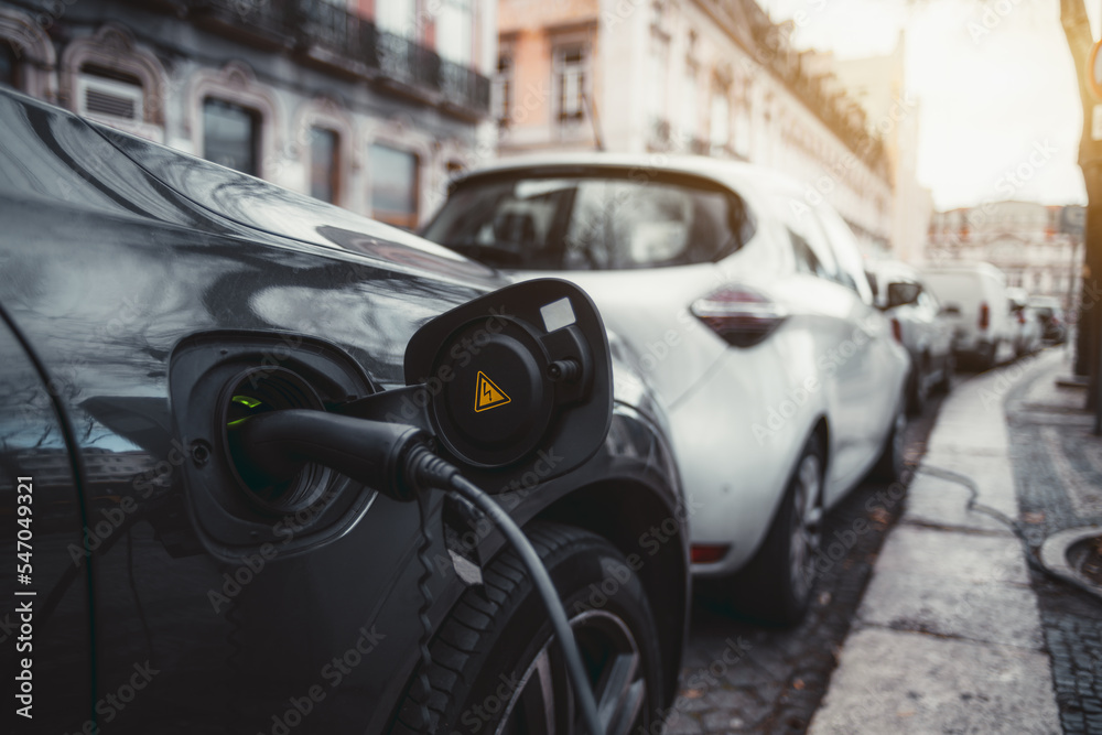 A wide-angle close-up shot of a thick black power cable inserted into an electric vehicle on the street; an electric car is charging via a black cord in a socket with a yellow alert sticker