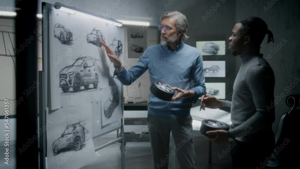 Two diverse automotive male designers holding small wheels, developing futuristic electric car design, standing near whiteboard with sketches. They working in car design development modern studio.