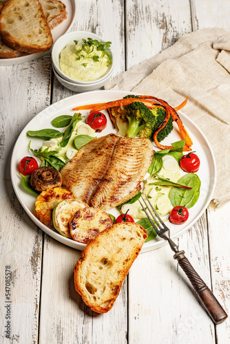 Fried sea fish and vegetables. Tilapia fillet with broccoli, cherry tomatoes and lettuce. Sea food on a white plate on white background. Vertical shot