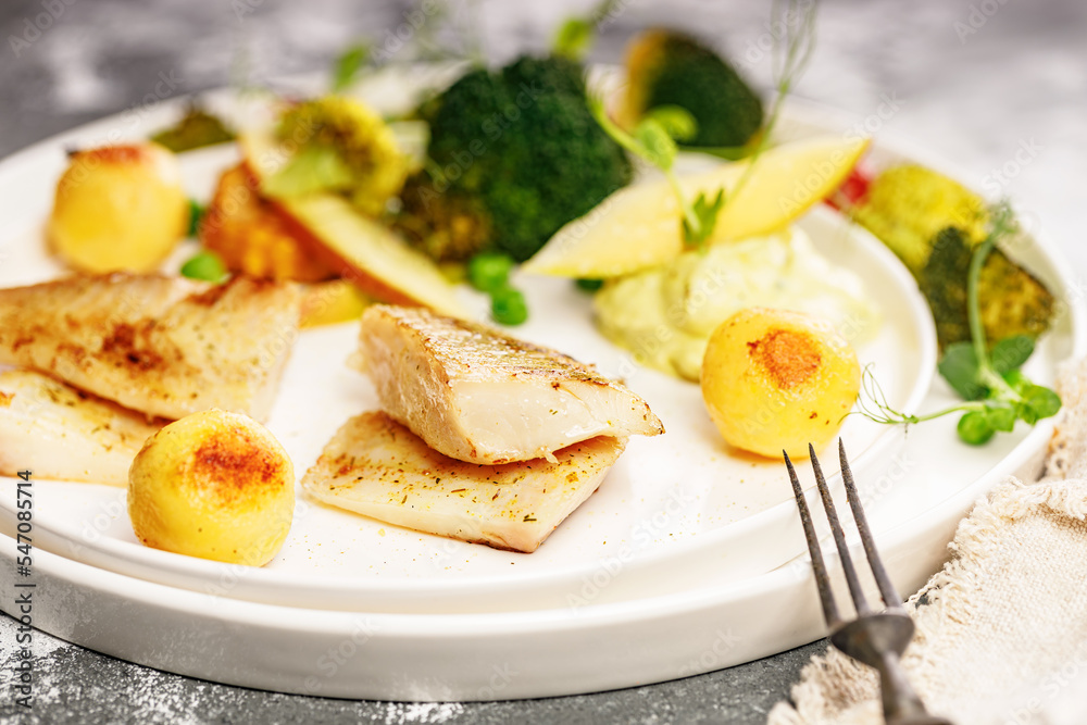 Close-up Baked sea fish fillet with vegetables. Tilapia fillet with broccoli, cherry tomatoes, fried potatoes, green peas and lettuce. Sea food on a white plate. Mediterranean Diet. Selective focus Close-up Baked sea fish fillet with vegetables. Tilapia fillet with broccoli, cherry tomatoes, fried potatoes, green peas and lettuce. Sea food on a white plate. Mediterranean Diet. Selective focus
