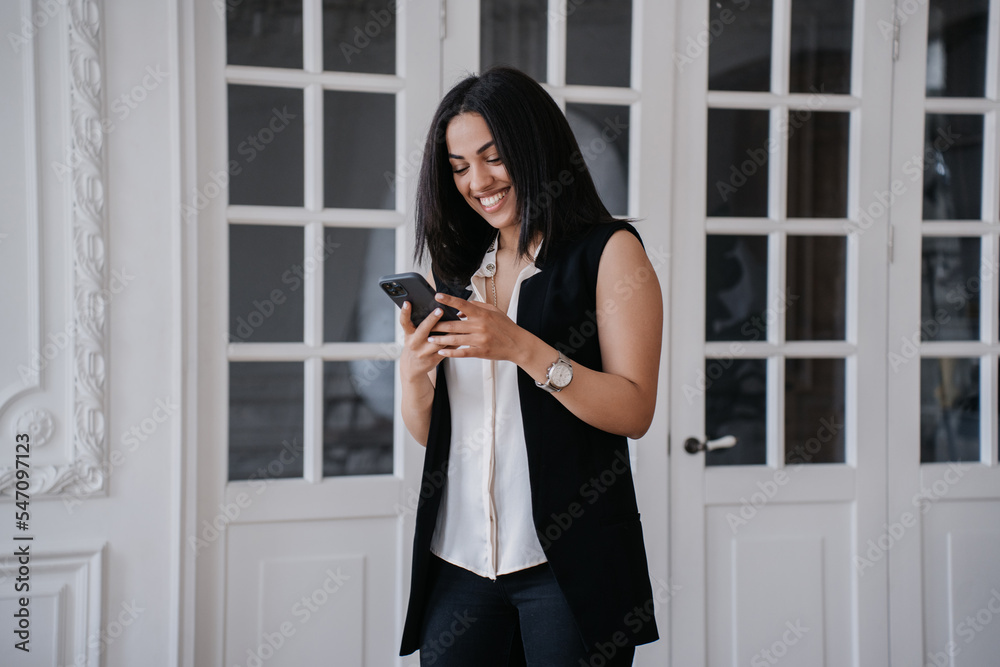 Adorable Brazilian girl holding phone, typing text message to boyfriend, smiles wide. Pretty African American young woman makes video call using cell phone at home. Happy businesswoman remote working.
