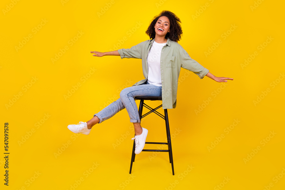 Full size photo of shiny young girl wavy hair sit stool spread hands smile wear stylish khaki clothes isolated on yellow color background