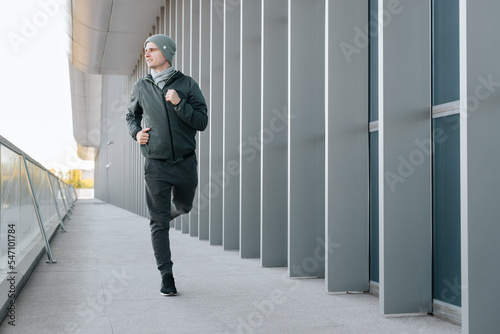 Young man in sportswear jogging in city of modern architecture. Smiling guy practicing sports in an urban environment, looking away