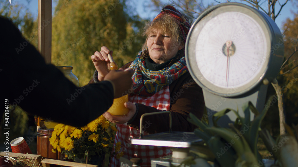 Obraz premium Elderly saleswoman stands at the stall, gives honey to taste. People pay for a jar of honey with cash. Local farmers market or autumn fair outdoors. Vegetarian and organic food. Agriculture.