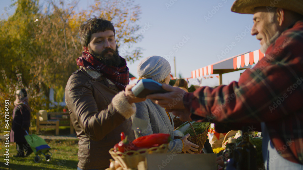 Couple shopping at local farmers market. Man contactless pays for purchases to farmer using smartphone. Autumn fair outdoors. Vegetarian and organic food. Agriculture. Point of sale system.