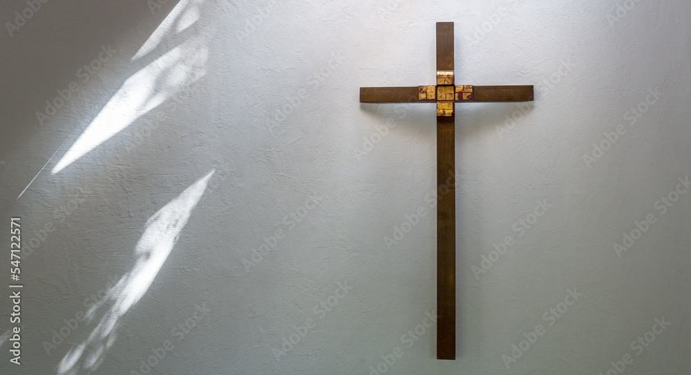 Christliches Kreuz aus braunem Holz mit Golddekor an einer weißen Wand, seitlich mit abstraktem Muster von einfallendem Sonnenlicht