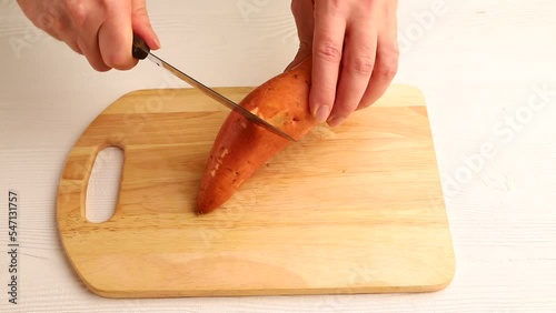 Male hand peels fresh batata with a knife on wooden kitchen board. Side view, close up.