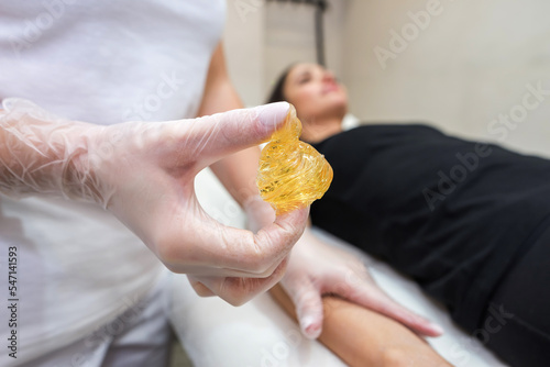 A female hand in a white glove holds a yellow sugar paste for depilation against the background of a lying client.