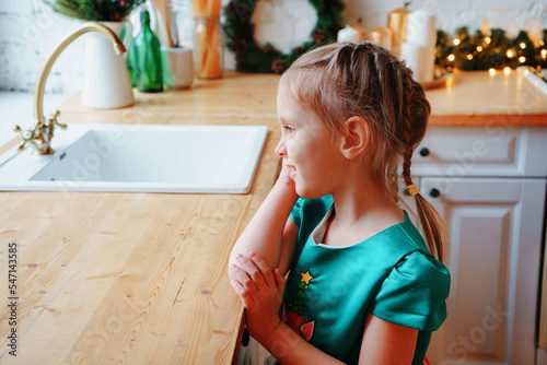 Adorable preschool girl wearing fancy bright green waiting for Christmas in the kitchen. Winter holidays concept.