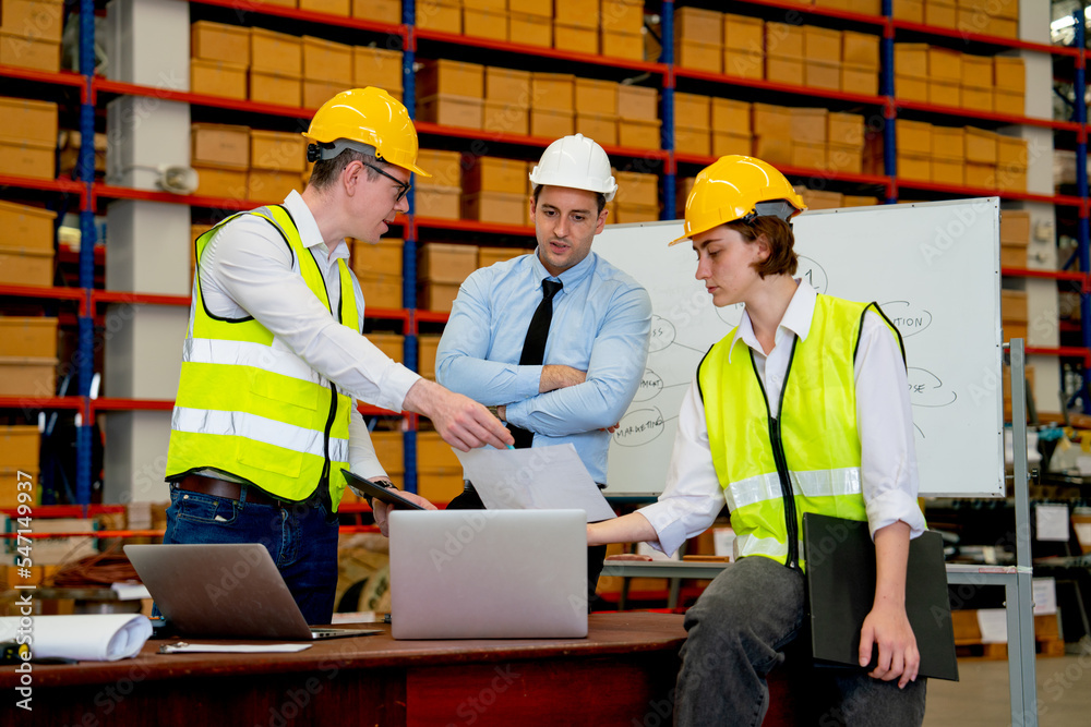 Three Caucasian warehouse workers discuss about the project using paper and laptop to support the explaination of their work. Three Caucasian warehouse workers discuss about the project using paper and laptop to support the explaination of their work.