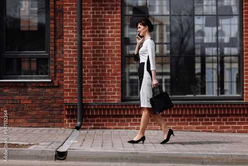 Happy lawyer businesswoman professional is walking outdoors talking on cell smart phone. Caucasian successful female is smiling wearing stylish white-black dress with black bag