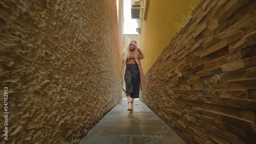 Young blonde-haired woman wearing fashionable clothes and holding purse defiles along narrow Venetian street between historical buildings photo