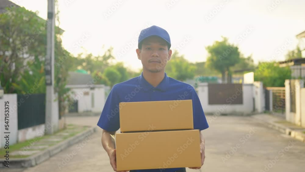 Handsome asian delivery man wearing blue uniform holding cardboard boxes in front of customer house. He smiling to camera feeling happy with his job. Handsome asian delivery man wearing blue uniform holding cardboard boxes in front of customer house. He smiling to camera feeling happy with his job.