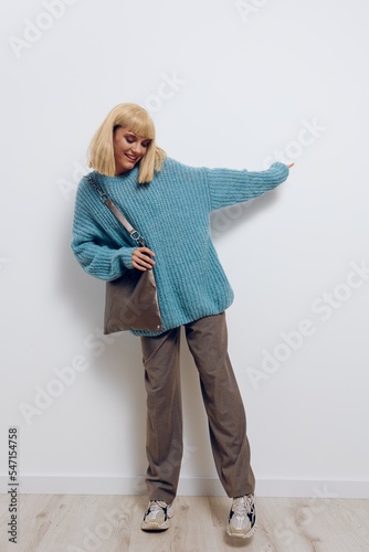 a full-length photo of a beautiful, happy, carefree stylish woman in a blue sweater and with a large bag, vigorously posing with her hand to the side