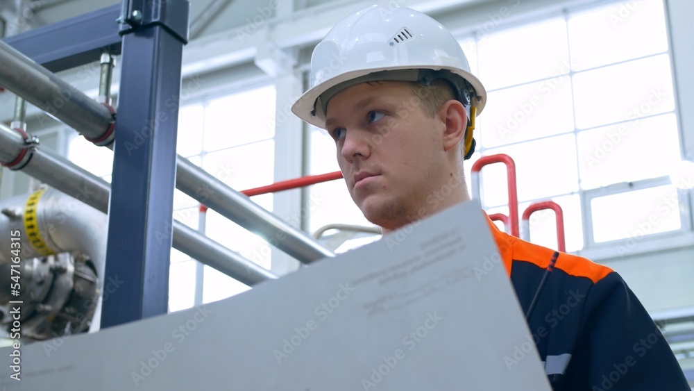A young engineer reads the technological scheme of the equipment at the plant. Work in heavy industry with mechanical equipment. Worker in overalls and a white work helmet at the workplace. A young engineer reads the technological scheme of the equipment at the plant. Work in heavy industry with mechanical equipment. Worker in overalls and a white work helmet at the workplace.
