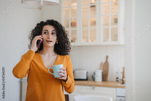 Surprised curly Hispanic woman in orange blouse talking by phone with blurry kitchen with excited face expression holds cup of coffee. Beautiful Caucasian housewife uses cellphone at home. Mockup.