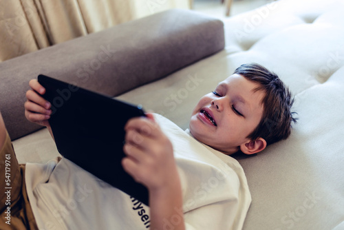 Little boy using digital tablet. Little boy with digital tablet sitting on sofa, on home interior background. Little boy reading an ebook. Growing up in a technology-based world.