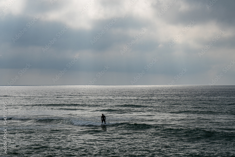 Young surfer man standing on his board submerged in the sea water under a cloudy sky about to rain and the water behind him illuminated with rays of the sunset sun coming out from between the clouds