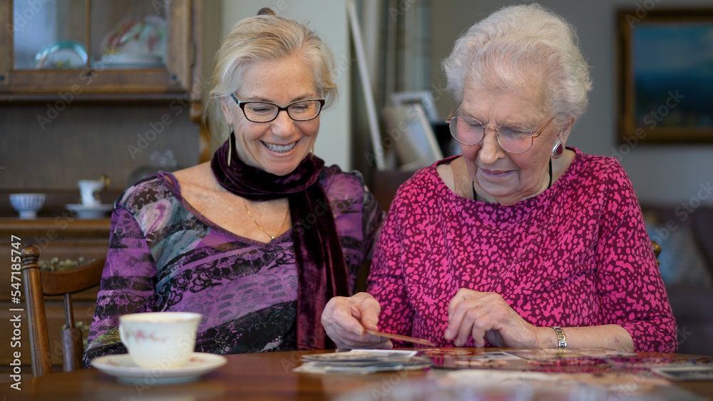 Senior elderly smiling happy woman looking at old photos and remembering memories with daughter at the dining room table. Senior elderly smiling happy woman looking at old photos and remembering memories with daughter at the dining room table.