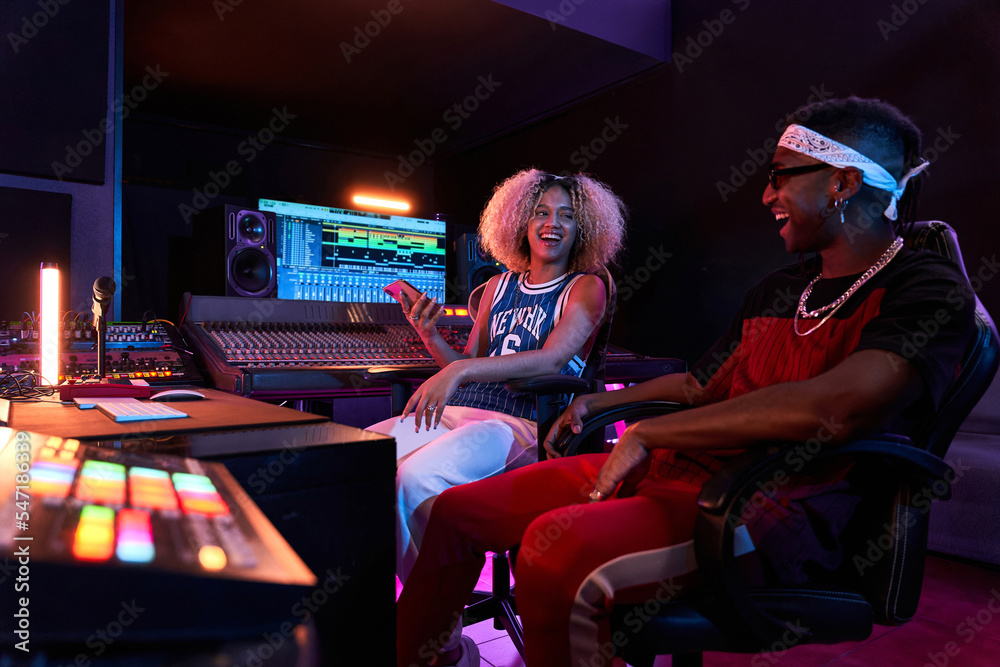 Cheerful black woman showing smartphone to partner sitting in recording studio Cheerful black woman showing smartphone to partner sitting in recording studio