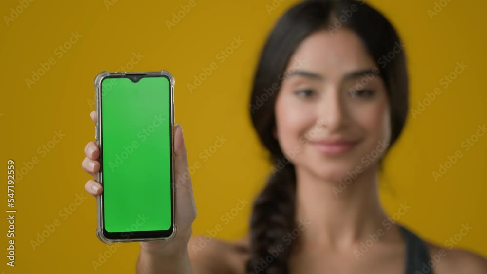 Indian ethnic woman lady girl lady in yellow studio background showing smartphone mobile telephone screen with green empty chroma key touchscreen technology cell message display advertise web service Indian ethnic woman lady girl lady in yellow studio background showing smartphone mobile telephone screen with green empty chroma key touchscreen technology cell message display advertise web service