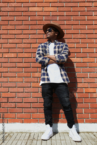 Young black man wearing wide brimmed fedora hat, sunglasses and checked shirt, posing over red brick wall background with copy space. Tanzania born hipster guy leaning on the wall.