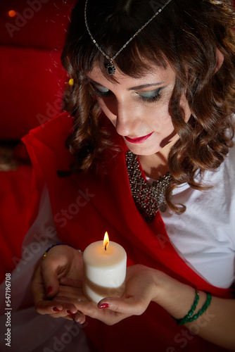 Beautiful arabian girl with candles in red room full of rich fabrics and carpets in sultan harem. Photo shoot of woman an oriental style odalisque. Model poses in sari as a caring wife and hostess