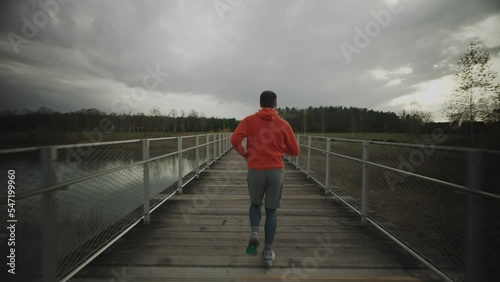 Male in orange hoodie running through national park across wooden bridge in wilderness in the cold fall weather. Sportsman does a fitness workout jogging on a timber footbridge in the countryside. 