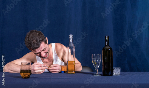 Lonely drunk man in homely white T-shirt sits at table and rips off wedding ring from his finger. In front of him are bottle of alcohol and glasses. Alcoholism as disease. photo