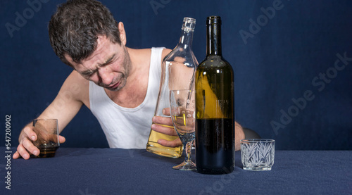 Lonely drunk man in homemade white T-shirt sits at table with his head down. In his hands he holds glass and bottle of vodka. Alcoholism as disease. photo